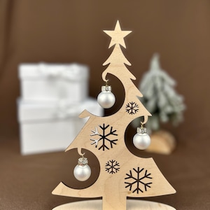 May include: A wooden Christmas tree decoration with a star on top and snowflake cutouts. It features three small, silver-colored ornaments. The base reads "Frohe Weihnachten".