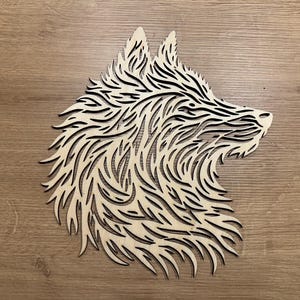 Puede incluir: Silueta de cabeza de lobo de madera, cortada con láser con detalles intrincados. El diseño presenta líneas superpuestas y fluidas que crean un efecto texturizado. La obra de arte se muestra sobre una superficie de madera, mostrando la veta natural.