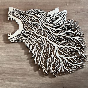 Puede incluir: Una pieza de arte mural de madera que representa la cabeza de un lobo con la boca abierta. El diseño presenta intrincados cortes en capas que crean un efecto texturizado y tridimensional. El pelaje y las características del lobo están estilizados con una estética natural y orgánica.