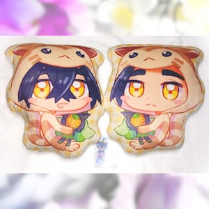 Puede incluir: Dos almohadas de felpa con un personaje de dibujos animados con cabello negro, que lleva un disfraz de gato con capucha de color marrón claro y sostiene un pequeño personaje de felpa verde. Las almohadas están sobre una superficie blanca.