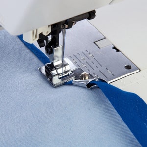 Puede incluir: Primer plano de una aguja y un pie de máquina de coser, cosiendo una tela azul con una tela blanca debajo. El pie es un tipo especial de pie para coser un ribete.