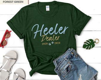 May include: Forest green t-shirt with the text "Heeler Dealer" in blue and gold lettering. The text "2020" and "2024" are also included in gold lettering, along with two paw prints.
