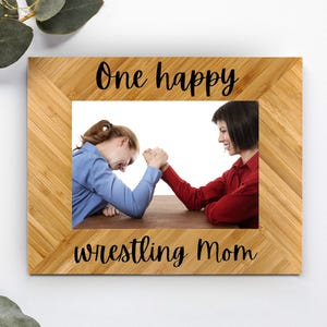 May include: A wooden picture frame with a light brown finish. The frame has a white mat and a photo of two people arm wrestling. The text "One happy wrestling Mom" is printed on the frame.