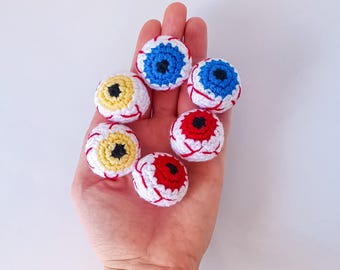 Könnte beinhalten: Eine Sammlung von sechs gehäkelten Augen in verschiedenen Farben, darunter Rot, Blau und Gelb. Jedes Auge hat eine schwarze Pupille und eine weiße Sklera.