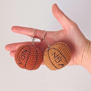 Puede incluir: Dos llaveros de baloncesto de crochet con la palabra "NBA" en ellos. Los llaveros son de color marrón y naranja y tienen costuras negras.