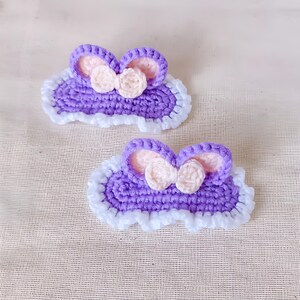 May include: Two handmade crochet hair clips. Each clip features a purple base with white trim, pink inner ears, and a cream-colored bow. The clips are designed in a cloud shape.
