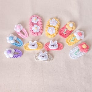 May include: A collection of 11 crocheted hair clips in various colours and designs. The clips feature flowers, bunnies, and other cute motifs.