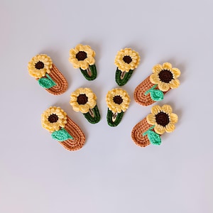 May include: A selection of handmade crochet sunflower hair clips. Each clip features a yellow sunflower with a brown centre and a green leaf. The clips are attached to brown and green bases, creating a charming and summery accessory.