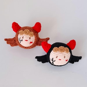 Puede incluir: Dos muñecos de ganchillo hechos a mano, uno marrón y otro negro, cada uno con cuernos rojos, alas y una cara con una cruz y una raya. Los muñecos tienen una cabeza redonda con una cara beige claro, una boca roja y una corona marrón.