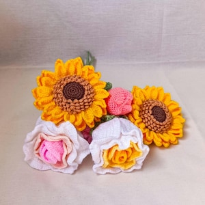 May include: A crocheted bouquet of flowers, including two sunflowers, a pink rose, and two white roses with yellow centers.