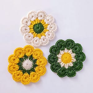 May include: Three crocheted flower coasters in white, yellow, and green. The coasters are made with a circular design and have a center circle with a smaller circle inside.