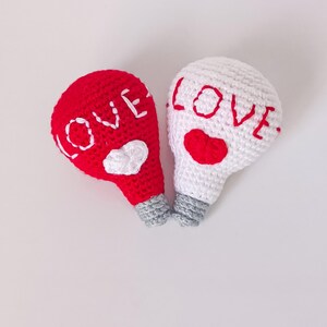 Puede incluir: Dos adornos de bombilla de crochet, uno rojo y otro blanco, con la palabra "LOVE" bordada en cada uno. La bombilla roja tiene un pequeño corazón blanco.