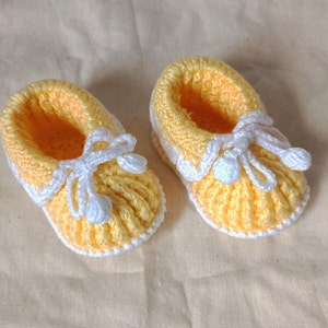 Könnte beinhalten: Ein Paar gelbe gehäkelte Babyschuhe mit weißen Schnürsenkeln und Bommeln.
