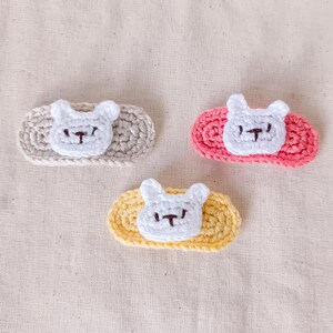 May include: Three handmade crochet hair clips. Each clip features a white bunny face with a black embroidered nose and mouth. The clips are set on oval bases in light gray, pink, and yellow. The background is a light beige fabric.