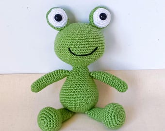 May include: A handmade green crochet frog doll with large, round eyes and a smiling mouth. The frog has outstretched arms and legs, and is sitting. The eyes are white with black pupils. The frog is made of a textured yarn.
