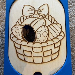 Op de afbeelding: Houten paaspuzzel met een blauw frame. De puzzel toont een mand gevuld met versierde paaseieren en een strik. Het ontwerp is in het hout geëtst. De tekst "LES CREATIONS EMMA 05" is zichtbaar.