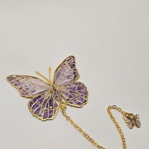 Puede incluir: Un objeto decorativo en forma de mariposa con un contorno dorado y un interior con diseños morados y blancos. Un pequeño dije de mariposa está unido por una cadena dorada. Probablemente un marcapáginas o un accesorio decorativo.