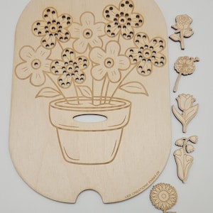 Pode incluir: Quebra-cabeça de flores de madeira com um design de flores em vaso e recortes de flores separados. A base do quebra-cabeça, marrom claro, tem detalhes gravados. Formas de flores adicionais incluem uma rosa, uma tulipa e um girassol. O texto "LES CREATIONS EMMA 06" é visível.