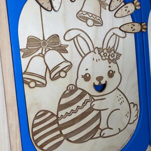 Peut inclure: Œuvre d'art en bois sur le thème de Pâques représentant un lapin souriant tenant un œuf rayé. Carottes, cloches et nœuds sont découpés au laser et fixés. L'œuvre est encadrée de bleu, créant une scène printanière joyeuse.