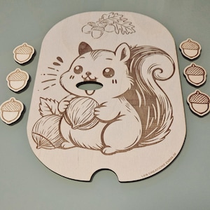 Peut inclure: Puzzle en bois sur le thème de l'écureuil, avec un écureuil de dessin animé tenant une noix, entouré de pièces en forme de gland. Le puzzle comporte une découpe pour la bouche de l'écureuil et est en bois clair. Des feuilles de chêne et des glands décoratifs sont également présents.