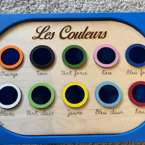 Può includere: Giocattolo didattico in legno con cornice blu. La tavola presenta dieci cerchi colorati etichettati in francese: Orange, Noir, Vert foncé, Rose, Bleu foncé, Blanc, Vert clair, Jaune, Bleu clair e Rouge.