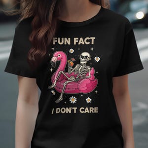 Könnte beinhalten: Schwarzes T-Shirt mit einem Skelett in Sonnenbrille auf einem rosa Flamingo-Schwimmring. Der Text lautet "FUN FACT I DON'T CARE", mit Gänseblümchen und Sternen. Ein humorvolles, lässiges Design.