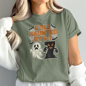 Może przedstawiać: Zielona koszulka z motywem Halloween. Projekt zawiera napis "STILL HAUNTED BY 6-7" w kolorze pomarańczowym, a także kreskówkowego ducha i czarną, zacienioną postać. Nietoperze i pajęczyny dopełniają upiorny design.