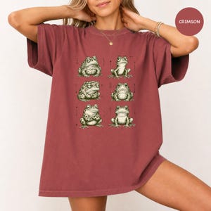 May include: A crimson short-sleeve t-shirt featuring a graphic print of six distinct green frogs arranged in two columns. Each frog is illustrated in a detailed, sketch-like style with varying poses and expressions, set against the solid fabric.