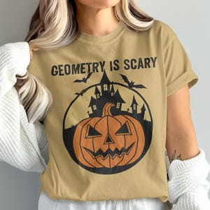 Może przedstawiać: Beżowa koszulka z napisem "GEOMETRY IS SCARY" nad czarną okrągłą grafiką. Grafika przedstawia dynię, nawiedzony dom i nietoperze, wszystko w kolorze czarnym i pomarańczowym. Koszulka jest w stylu casual z krótkim rękawem.