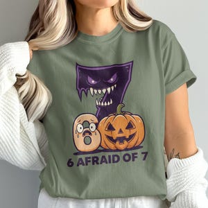 Może przedstawiać: Szałwiowa zielona koszulka z motywem Halloween. Projekt zawiera fioletowego potwora, liczbę 7, dynię i liczbę 6 z przerażoną miną. Tekst "6 AFRAID OF 7" jest wydrukowany pod liczbami i dynią.