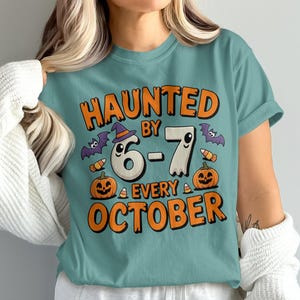 Może przedstawiać: Turkusowa koszulka z napisem "HAUNTED BY 6-7 EVERY OCTOBER" w kolorze pomarańczowym. Projekt zawiera ducha w kapeluszu czarownicy, nietoperze, cukierki i dynie. Koszulka graficzna z motywem Halloween.