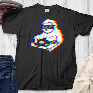 Puede incluir: Camiseta negra con un perezoso usando gafas de sol y una mesa de mezclas. El diseño del perezoso y la mesa de mezclas tiene un efecto 3D con contornos rojos, azules y amarillos. La camiseta es de la marca Gildan.