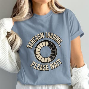 May include: A dusty blue short-sleeved t-shirt featuring a circular loading bar graphic in the center. The text "SARCASM LOADING..." is arched above the graphic, and "PLEASE WAIT" is arched below, both in a distressed, off-white font.