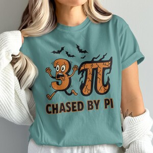 Może przedstawiać: Turkusowa koszulka z zabawną grafiką. Projekt przedstawia liczbę 3 uciekającą przed symbolem Pi, z nietoperzami nad nią i tekstem "CHASED BY PI" poniżej. Grafika jest w odcieniach pomarańczu i brązu.