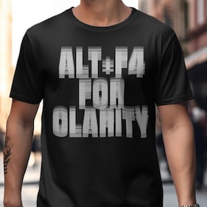 Puede incluir: Camiseta negra con el texto "ALT+F4 FOR CLARITY" en fuente blanca superpuesta. La camiseta de manga corta parece ser de un material suave. El fondo está borroso, sugiriendo un entorno exterior.