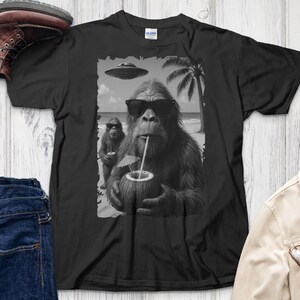 May include: Black t-shirt featuring a Bigfoot graphic wearing sunglasses and drinking from a coconut on a beach. A smaller Bigfoot is in the background. A UFO and palm tree are also in the graphic.