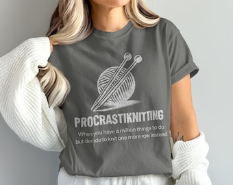 Op de afbeelding: Grijs t-shirt met een witte afbeelding van breinaalden en garen, en de tekst "PROCRASTIKNITTING". Het shirt heeft ook de tekst "When you have a million things to do but decide to knit one more row instead."