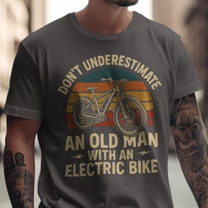 Puede incluir: Camiseta gris oscuro con el texto "DON'T UNDERESTIMATE AN OLD MAN WITH AN ELECTRIC BIKE" sobre un gráfico de una bicicleta eléctrica. El diseño presenta un fondo retro de puesta de sol.