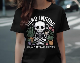 Pode incluir: Camiseta preta com um desenho de esqueleto segurando uma xícara de café, rodeado por plantas em vasos. O texto diz "DEAD INSIDE BUT MY PLANTS ARE THRIVING". O design é em estilo cartoon com cores pastel.