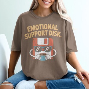 May include: Brown t-shirt with the text "EMOTIONAL SUPPORT DISK." The graphic features a floppy disk character wearing sunglasses and holding a coffee cup. The shirt is a comfortable fit.