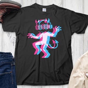 May include: Black t-shirt featuring a glitch-effect devil character in pink, blue, and white. The devil has horns, a tail, and headphones. The shirt is displayed with jeans, a jacket, and boots.