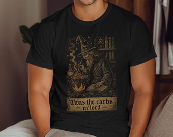 Op de afbeelding: Zwart t-shirt met een illustratie van een tovenaar die pasta kookt boven een vuur, met de tekst "Twas the carbs, m'lord". De tovenaar draagt een hoed en is omringd door boeken. Een humoristisch ontwerp.