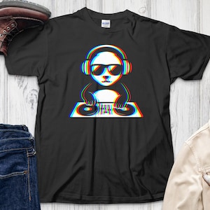 Puede incluir: Camiseta negra con un diseño de panda DJ con efecto 3D. El panda lleva gafas de sol y auriculares, y está situado detrás de una mesa de mezclas. El diseño utiliza colores rojo, azul y blanco. La camiseta se muestra con vaqueros, una chaqueta y botas.