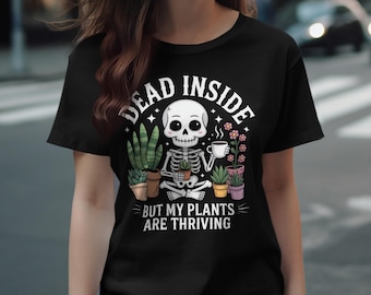 Pode incluir: Camiseta preta com um desenho de esqueleto de desenho animado segurando uma xícara de café, rodeado por plantas em vasos. O texto diz "DEAD INSIDE BUT MY PLANTS ARE THRIVING". O design incorpora estrelas e flores, oferecendo um estilo peculiar.