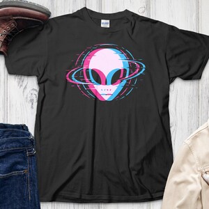 Pode incluir: Camiseta preta com um design de cabeça alienígena rosa, azul e branco com um anel semelhante a um planeta. A imagem inclui jeans azuis, uma bota marrom e uma jaqueta bege com óculos.