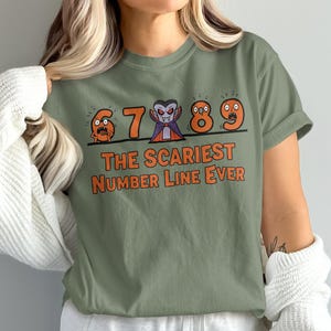 Może przedstawiać: Zielona koszulka z motywem Halloween. Projekt przedstawia liczby 6, 7, 8 i 9, każda z przerażającą twarzą, oraz liczbę 7 przypominającą wampira. Tekst brzmi: "NAJSTRASZNIEJSZA LINIA LICZBOWA W HISTORII."