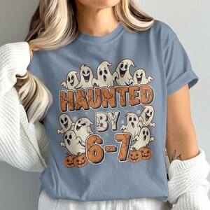 Może przedstawiać: Jasnoniebieska koszulka z motywem Halloween. Projekt przedstawia kreskówkowe duszki i napis "HAUNTED BY 6-7" w kolorze pomarańczowym i brązowym, z dyniami. Koszulka jest w stylu casual, z krótkim rękawem.