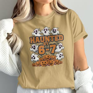Może przedstawiać: Musztardowa koszulka z grafiką o tematyce Halloween. Projekt przedstawia kreskówkowe duchy, dynie i napis "HAUNTED BY 6-7" w kolorze pomarańczowym i czarnym. Koszulka wykonana jest z miękkiego materiału.