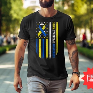 Op de afbeelding: Zwart T-shirt met een blauw en geel lintontwerp op een witte achtergrond. Het lint is een symbool van Down Syndrome Awareness. De tekst "Down Syndrome Awareness" staat verticaal aan de rechterkant van het lint.