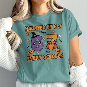 Może przedstawiać: Turkusowa koszulka z napisem "HAUNTED BY 6-7 EVERY OCTOBER". Projekt przedstawia fioletową cyfrę 6 w kapeluszu czarownicy i pomarańczową cyfrę 7 w pelerynie, a także dynie, nietoperze i cukierki.
